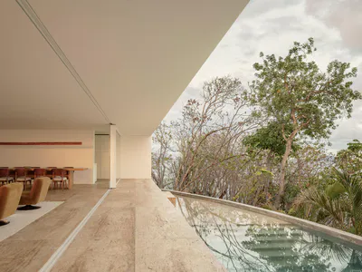 Modern open-plan interior with travertine floors, brown velvet swivel chairs, long wooden dining table, and an infinity pool overlooking a lush tropical cliffside and ocean.