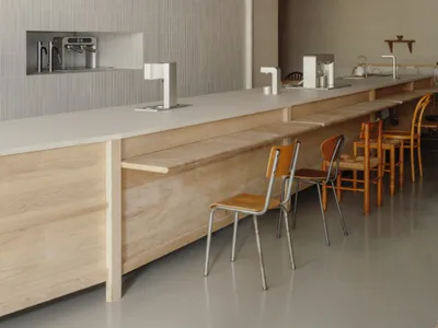 Modern minimalist cafe interior with a long light-wood plywood counter, metal stools with orange seats, exposed silver HVAC ducts on a concrete ceiling, and a recessed stainless steel coffee station.