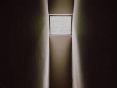 Looking up a narrow concrete staircase with a handrail on the left and a high rectangular window at the top allowing diffused daylight.