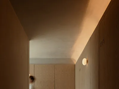 A long, minimalist hotel hallway with light oak wood paneling on walls and doors, round wall sconces emitting warm light, and a person captured in motion blur walking from a connecting corridor.