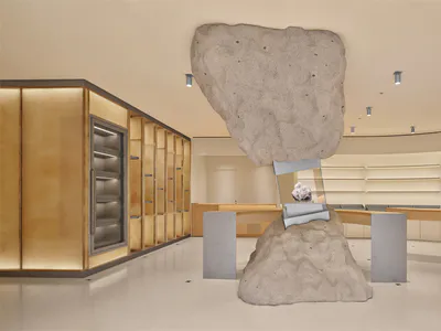 Modern luxury retail interior featuring a massive floating boulder sculpture suspended over a mineral display case on a raw stone base, with curved wooden counters and illuminated display cabinets in the background.