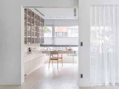 View into a bright minimalist home office featuring light wood built-in cabinetry, a simple wooden desk and chair, large windows with roller blinds, and white sheer curtains on the right.