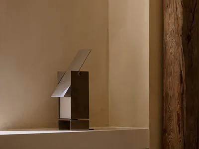 Modern stainless steel abstract sculpture placed on a white plaster windowsill next to a textured wooden beam in a minimalist interior space.