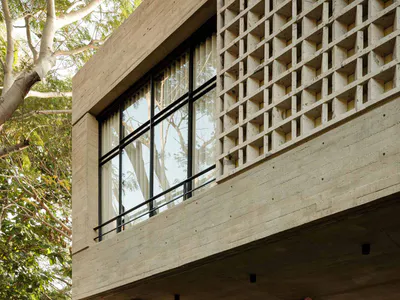 Modern tropical house exterior featuring exposed concrete structure, large black-framed windows, built-in entertainment niche, and an open-air patio furnished with natural wicker lounge chairs and sofas surrounded by abundant tropical plants.