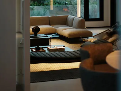 Modern minimalist living room interior featuring a beige linen sectional sofa, low black coffee tables, a woven daybed, and tall floor lamps, with large windows overlooking a dense green forest.
