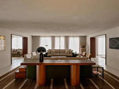 A spacious modern minimalist living room featuring a large round walnut dining table in the foreground, a green sectional sofa, a beige sofa in the background, and large windows with sheer curtains allowing soft daylight to fill the space.