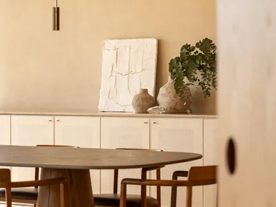 Modern minimalist dining room featuring a round oak pedestal table, wooden chairs with brown leather seats, a sculptural white textured artwork, ceramic vases with eucalyptus, and a two-tier white pendant light against a textured beige wall.