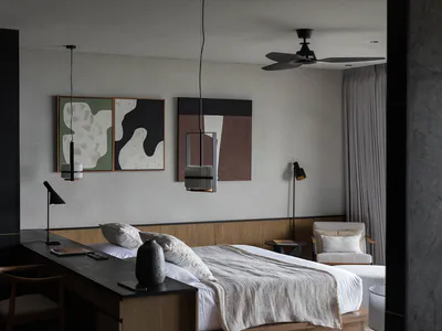 Modern minimalist bedroom interior with a wooden platform bed, dark wood desk and chair, abstract green and brown wall art, black pendant lights, and a black ceiling fan.