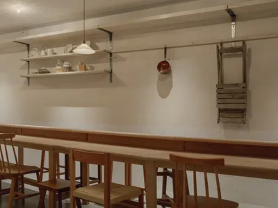 Long wooden communal dining table with matching oak chairs, floating shelves with dishware, a hanging copper pot, and a wooden ladder on a white tiled wall.