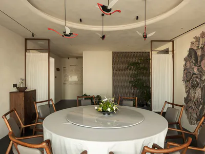Modern Chinese-style dining room with a large round marble lazy Susan table, wooden dining chairs with grey upholstery, dramatic Tang dynasty mural on the right wall, and artistic red and white ceiling fans hanging from a curved ceiling.