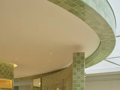 Modern boutique interior featuring curved walls covered in glossy green ceramic tiles, a minimalist clothing rack with a yellow shirt, and a cylindrical shelving unit with warm backlighting.