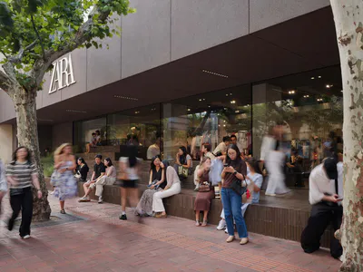 現代Zara店面前熱鬧的都市廣場，大面玻璃窗、洞石階梯作為座椅，人們在陽光下的大樹旁休憩。