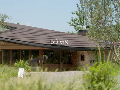 BG Café 外觀，呈現長弧形棕色木屋頂、大面玻璃窗、石材砌牆，以及藍天下環繞的樹木與植栽。