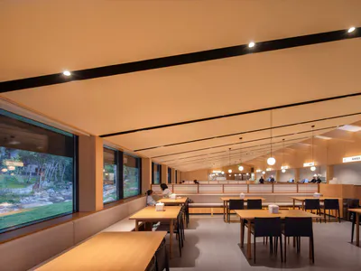 Modern minimalist restaurant interior featuring extensive light wood finishes, built-in banquette seating along large windows, wooden dining tables with black chairs, and warm ambient lighting.