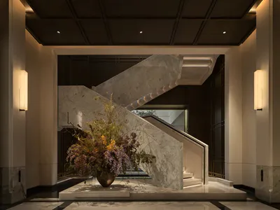 View of a luxurious hotel lobby featuring a dramatic white marble grand staircase with integrated lighting, a large bronze vase with an elaborate dried floral arrangement in autumn tones, dark coffered ceiling, and polished marble floor with black inlays.