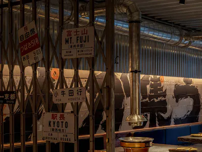 日式火鍋餐廳內部，工業風金屬伸縮門上掛滿復古火車站牌，藍色沙發座椅與中央銅製火鍋置於深色石材吧台。