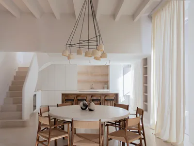 Modern minimalist dining room featuring a large round travertine table surrounded by six wooden woven chairs, with a sculptural black pendant light above, white walls, a curved staircase on the left, and an open kitchen with wooden bar stools in the background.