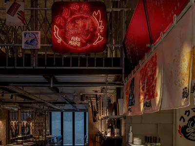 日式居酒屋室內空間，設有長型磁磚壽司吧台、木椅、清水模牆面、懸掛的暖簾，以及醒目的紅色霓虹鬼面招牌。