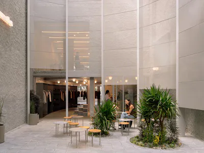 Modern retail atrium featuring textured concrete walls, large glass panels, indoor plants, and circular tables with people relaxing in a bright, open space.