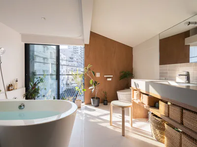Modern minimalist bathroom interior with white freestanding bathtub filled with water, wooden accent wall, multiple potted plants, wall-mounted faucet, woven storage baskets, and large glass doors opening to a city balcony.