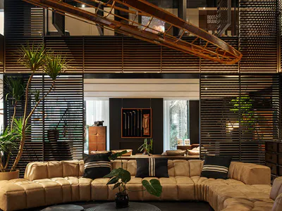 Modern double-height living room featuring a large curved tan leather sectional sofa, black and white striped pillows, round dark wood coffee tables, potted plants, and a striking wooden boat sculpture hanging from the ceiling with wooden slat walls in the background.