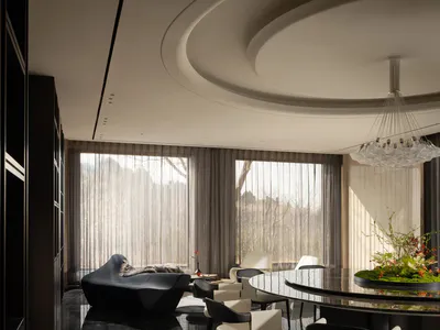 Modern luxury dining and living area featuring a large round black marble table, cream and black dining chairs, a sculptural black chaise lounge, dramatic curved ceiling with a large crystal chandelier, and floor-to-ceiling windows overlooking a forest view.
