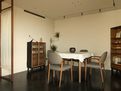 Modern minimalist dining room with a rectangular table covered in white linen, four grey upholstered wooden dining chairs, a tall dark wood display cabinet filled with ceramics, a matching tall bookshelf, potted plants, and a dark glossy floor under track lighting.