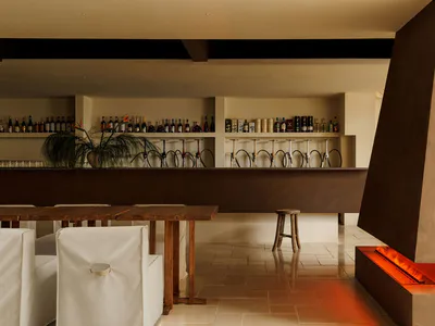 Modern bar interior with dark corten steel counter, backlit bottle shelves, wooden tables, white upholstered chairs, and a glowing linear fire feature on the right.
