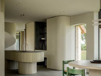 Modern minimalist dining area with curved oak kitchen island, moss green wooden chairs with woven seats, and a sculptural white rug on grey tiled floor.