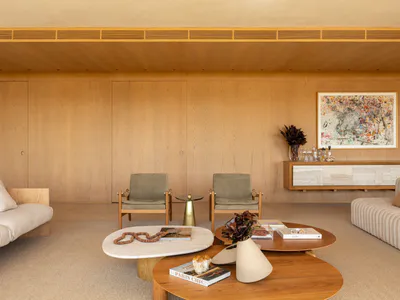 Modern minimalist living room featuring extensive oak wood paneling, two green upholstered armchairs, a white sofa, and three nesting coffee tables made of marble and wood, with colorful abstract art on the wall.