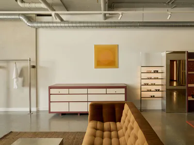 Modern boutique interior with a mustard tufted sofa in the foreground, a pink-trimmed white credenza, illuminated eyewear display shelves, and a large ochre circular artwork on a white wall with exposed silver ductwork.