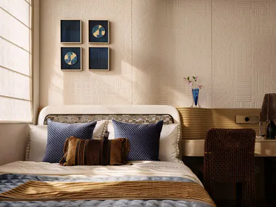 Modern neutral-toned bedroom with a bed dressed in white and blue bedding, multiple blue and brown decorative pillows, a mosaic headboard, textured beige wall panels, four pieces of blue wall art, a wooden desk with a woven brown chair, and a blue glass vase with flowers.