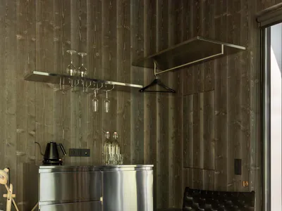 Modern hotel room corner with dark wood walls, stainless steel minibar, floating glass shelves with wine glasses, black leather woven chair, and a tree stump side table.
