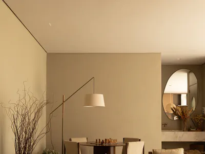 Modern minimalist dining room featuring a round walnut chess table with upholstered chairs, a dramatic floor lamp, large terracotta vases with dried branches, and an oval mirror reflecting another part of the home.