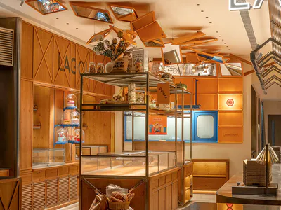 現代麵包店內部，展示暖木質櫃體、玻璃展示櫃，以及懸掛橘色鏡面的創意天花裝置，反射出溫暖光線。