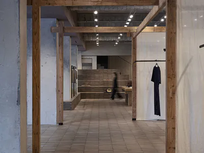 Long perspective view of a minimalist boutique interior featuring exposed concrete walls, large wooden structural beams, tiled floor, and clothing displays under track lighting.