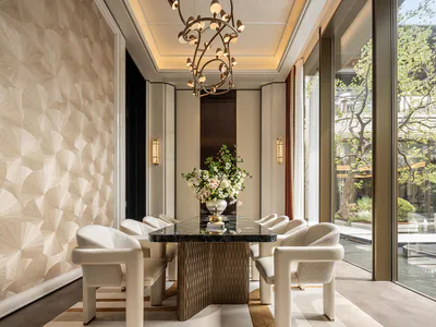 Modern luxury dining room with black marble table, white upholstered dining chairs, sculptural chandelier, textured beige wall panels, and large glass doors opening to a garden courtyard.