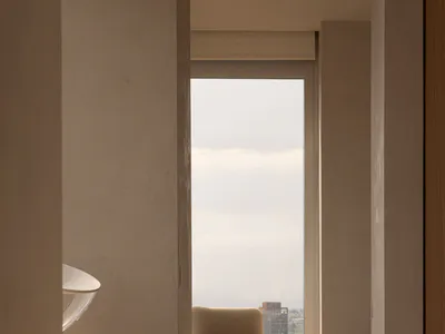 View from a doorway into a minimalist room featuring a beige upholstered oak armchair, a matching ottoman, and a large window overlooking a hazy city skyline with dramatic morning light casting shadows on light wood floors.