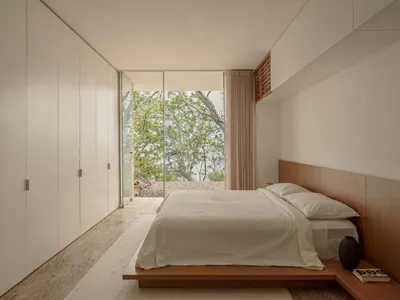 Modern minimalist bedroom with a low wooden platform bed, white linens, built-in wood headboard, white wardrobe, and large glass doors revealing green trees and a rocky ocean view.