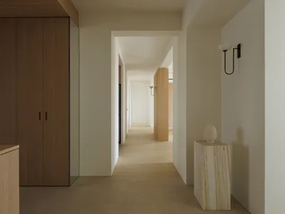 Long minimalist hallway featuring a travertine stone pedestal with a white ceramic vase, warm wood cabinetry, black wall sconce, and light oak flooring.