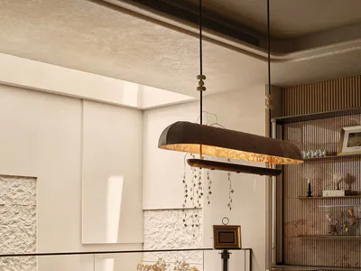 Modern home office featuring a large curved travertine desk with integrated shelving, a sculptural two-tier pendant light with hanging crystals, a textured stone wall, and built-in wooden shelving displaying decorative objects.