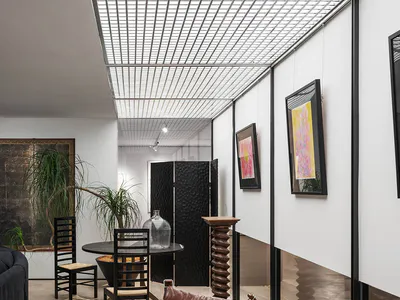Modern gallery-style living area featuring a brown leather sling chair, black dining chairs, potted plants, framed abstract art, and a book titled Seeds & Fruits on a side table.