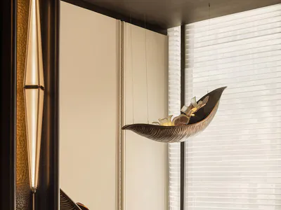 Modern luxury interior featuring a sculptural bronze leaf-shaped hanging lounge chair suspended from the ceiling and a matching floor version, with a tall pendant light on the left and large windows with sheer blinds in the background.