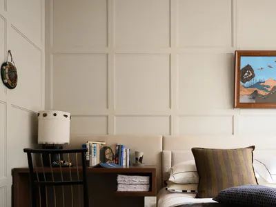 Modern neutral bedroom featuring a walnut desk, black spindle chair, upholstered bed with striped and textured pillows, table lamp, and framed landscape painting on a paneled wall.