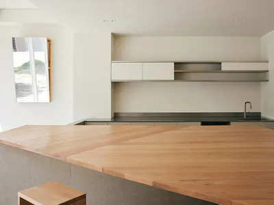 Modern minimalist kitchen featuring a large solid oak island with herringbone pattern, concrete base, white handleless cabinets, stainless steel floating shelves, and a stainless steel countertop with sink.