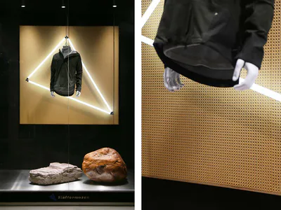 Modern retail display case with a black technical jacket suspended inside a glowing triangular light frame against a golden perforated metal panel, accompanied by two large natural stones on a stainless steel base.