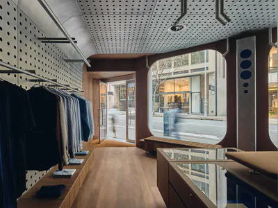 Interior view of a contemporary clothing boutique with walnut wood display fixtures, perforated metal ceiling, clothing racks, and large arched street-facing windows.