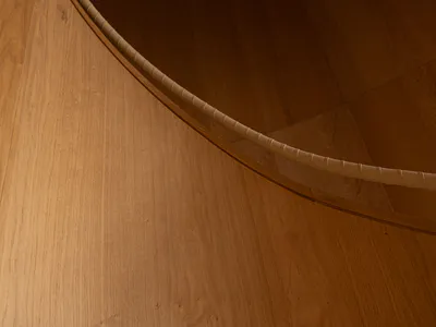 Close-up photograph of the curved edge of a thick solid oak round dining table positioned over a light oak wood plank floor, showing the smooth tabletop and ribbed side detail.