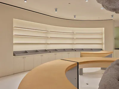 Modern minimalist retail interior featuring a large curved oak counter, built-in beige cabinetry with grey bins, and floating illuminated shelves under a curved ceiling with spotlights.