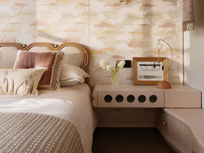 Cozy bedroom corner featuring a curved rattan headboard, layered neutral and blush pillows, textured abstract wallpaper in cream, pink and beige, and a floating night console with modern lighting.